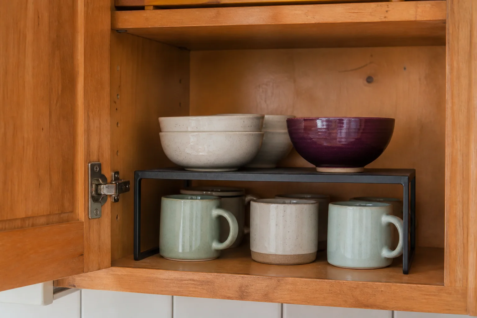 Metal shelf riser separating lightweight bowls and mugs inside a cabinet